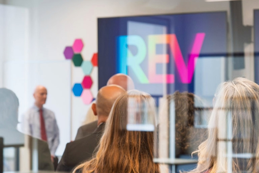 Audience listening to Philip Steenkamp, President of Royal Roads University, speaking in a meeting room with a presentation screen.