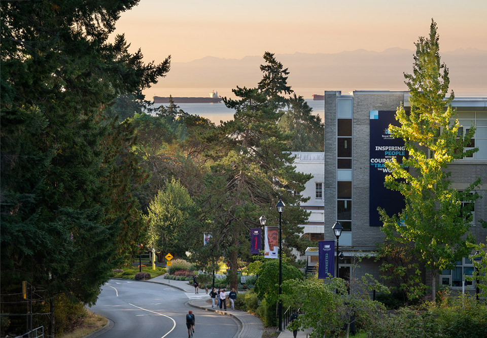 An aerial view of Royal Roads University, showing a winding road, tall trees, buildings and the Olympic Mountains in the backgroud.