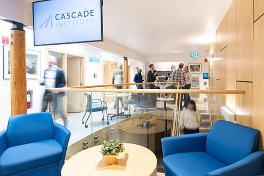 A room full of people talking in a group with a TV hanging from the ceiling reading "Cascade Institute"