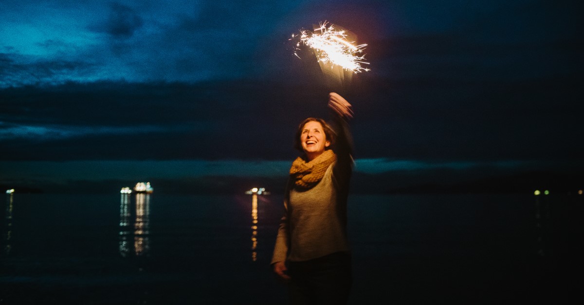 Cheryl Heykoop waving a sparkler in the night.  Distant boat lights reflect in the water behind her.