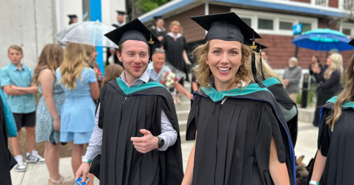 Two Royal Roads graduates walk by a crowded outdoor area during a convocation celebration.