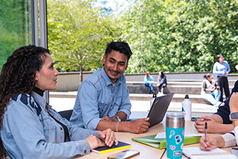 Two RR students sit at a table next to an open door. This image links to information for visiting and exchange students.