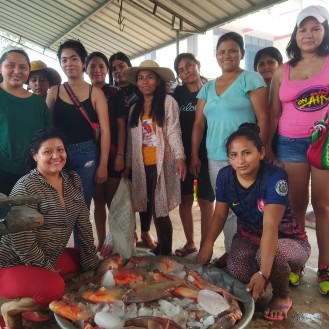 A group of women smiling and looking at the camera with a large container of fish on ice at their feet.