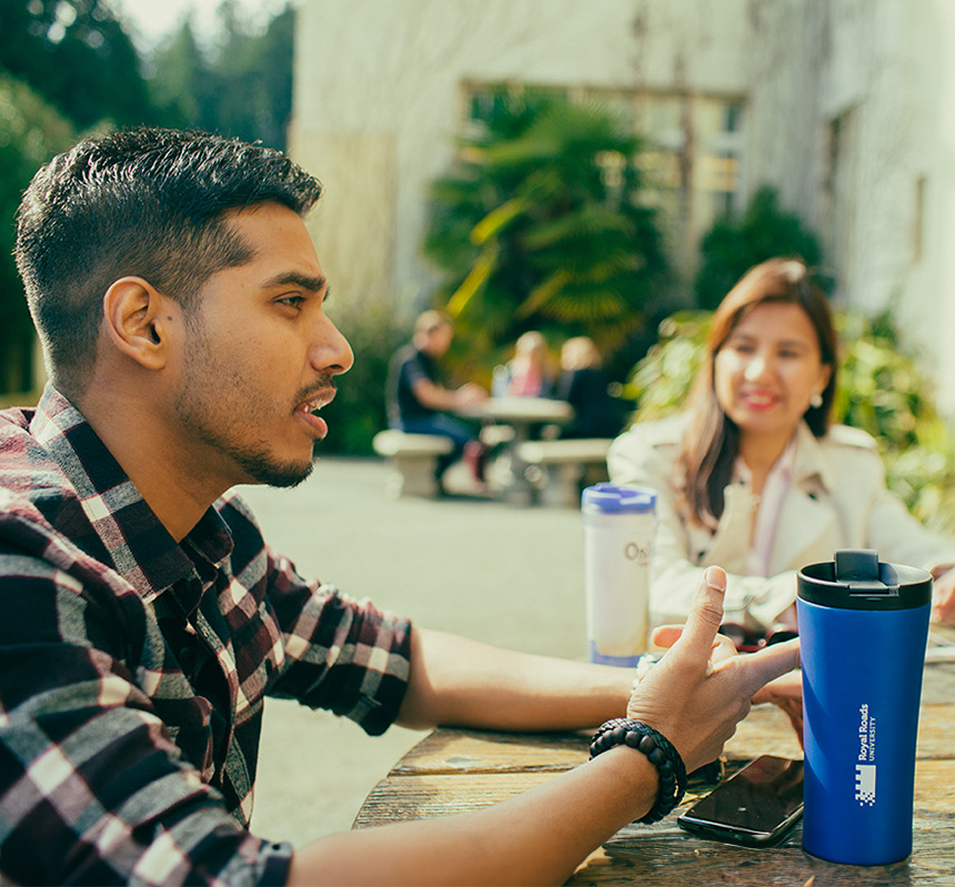 An RRU student sits at a picnic table with their peers on a sunny day with a blurred background of trees and buildings behind them