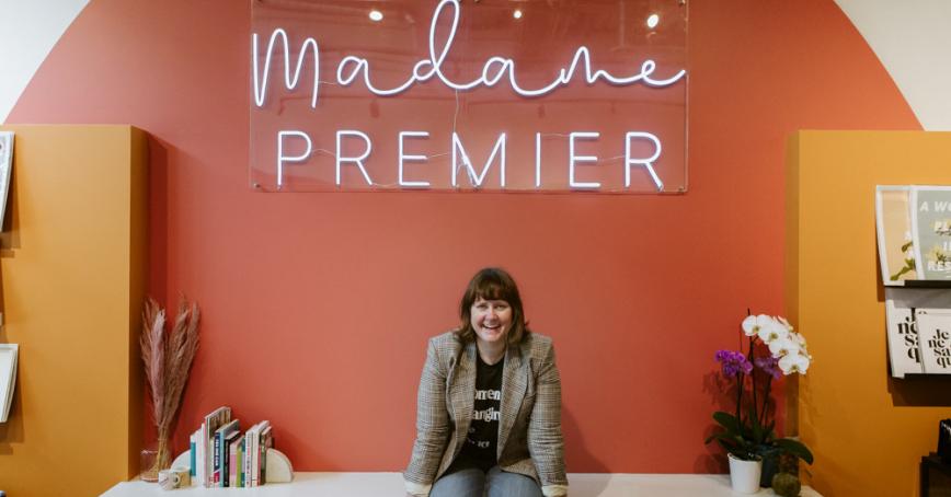 smiling person sits on a counter at a store. The words "Madame Premier" in neon white affixed to a sign above their head.