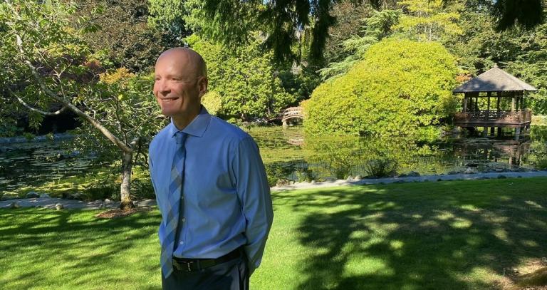 President Philip Steenkamp in the Japanese Gardens at Royal Roads University. He is smiling and looking  into the gardens. In the background is the pond, dappled light and bridge.