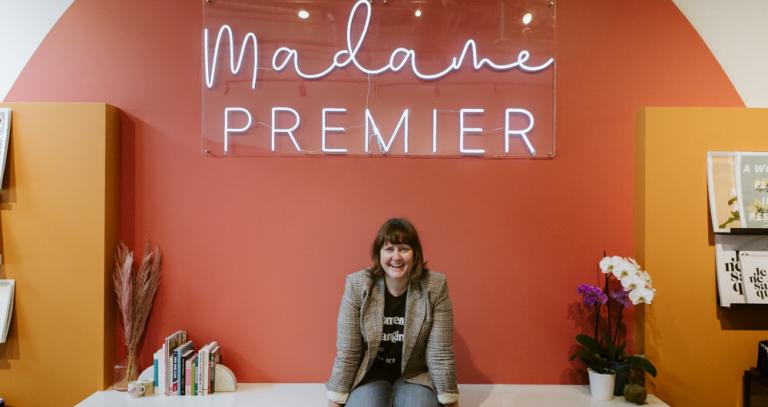 smiling person sits on a counter at a store. The words "Madame Premier" in neon white affixed to a sign above their head.