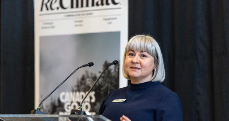 Woman with short grey hair and bangs standing at a podium with a sign saying Re,Climate on the wall behind her.