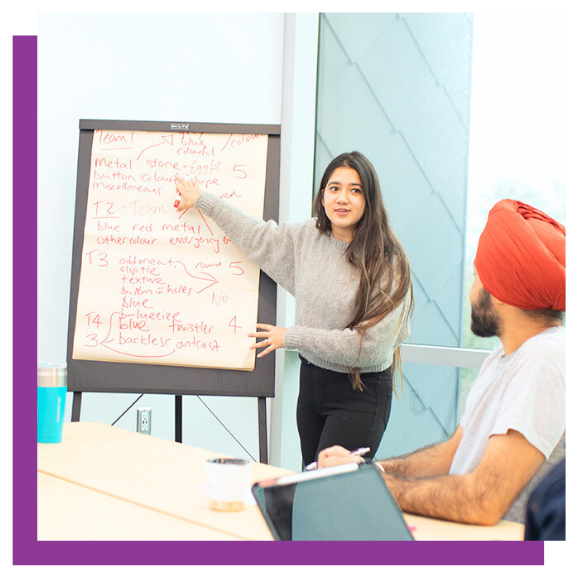 A student presenting notes on a flip chart to a seated colleague in a meeting room