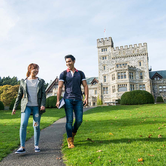 Two students walk down a grassy path in front of Hatley Castle under a blue bright sky