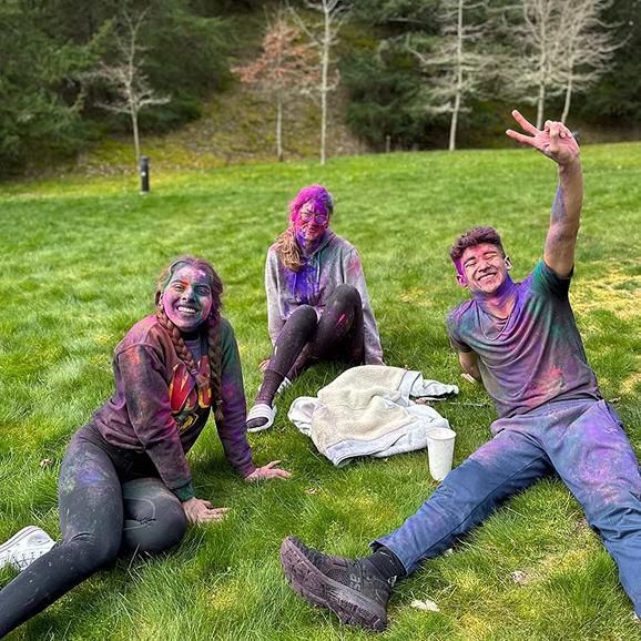 Three students covered in colorful powder celebrating Holi on the RRU campus lawn
