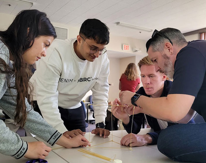 A group of BBA students huddle over a large table to assemble a tower made of pasta.