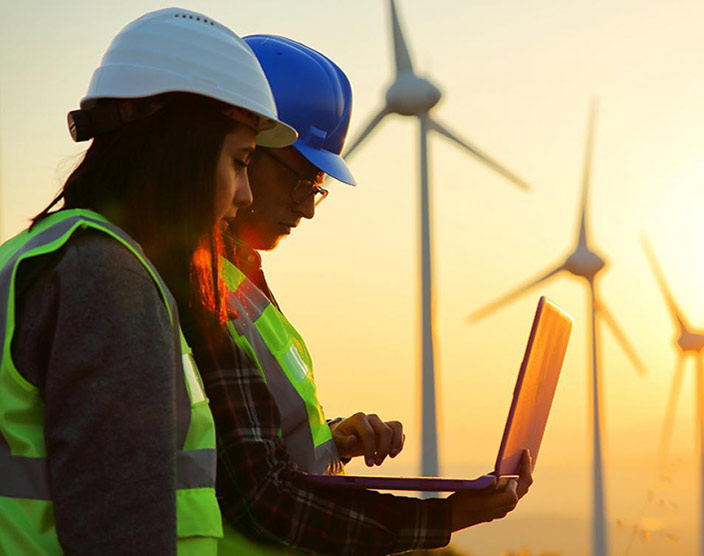 Two RRU students wearing yellow safety vests stand next to a row of wind turbines, look at a laptop