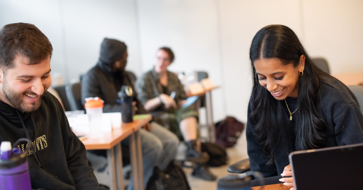 Two students in the classroom smile and look down at their work. In the background other students are chatting.