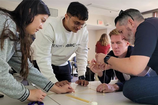 Four students lean over a table in a team-building exercise, holding spaghetti and building a tower