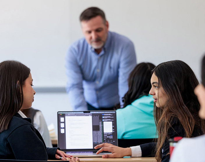 A professor leans over a desk while two of his students look toward each other with an open laptop in front of them.