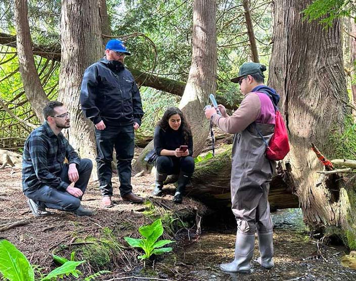 Four RRU students squat next to a large tree in a forested area as part of an outdoor learning experience.