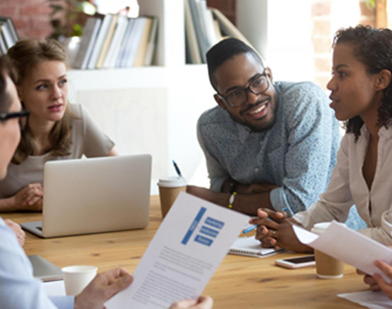 A group of students sit around a table with their laptops and paperwork while in conversation. This links to the Graduate Certificate in Management and Leadership page.