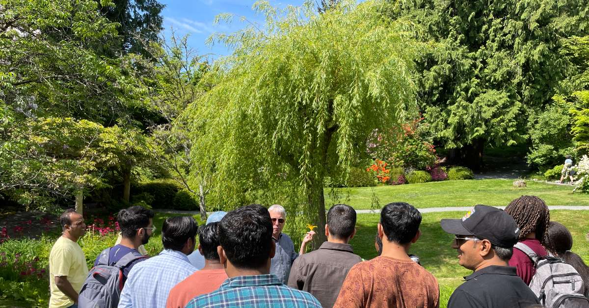 Students standing in a garden looking at teacher, who is holding up a flower.