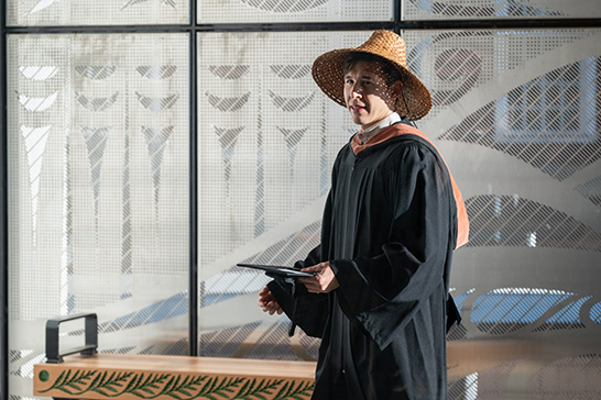 Person wearing academic regalia and a woven hat stands indoors, holding a tablet, with patterned glass panels in the background.