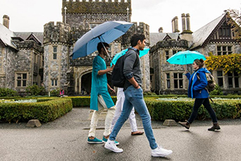 A group of international students holding umbrellas pass by Hatley Castle. This image links to information for international students.