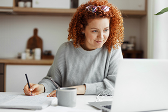 A woman sits at her desk looking at her laptop and taking notes as part of on online program. This image links to a page about the RealEstate Development Micro-credential