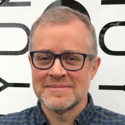 Rhys Lloyd smiling wearing dark rimmed glasses and a blue and black collared shirt.