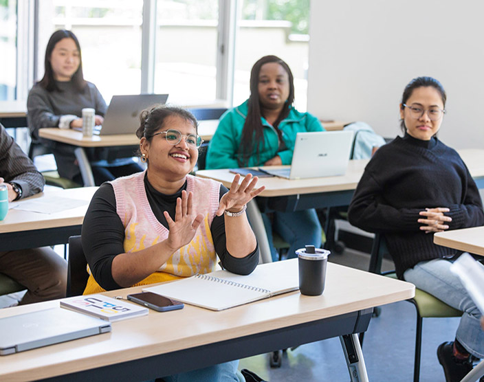 A student lifts her hands while speaking in a classroom, while her peers sit at their desks and look on in admiration.
