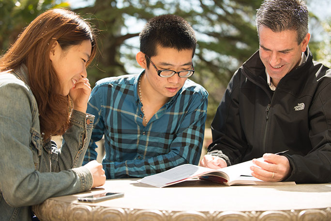 Three people sit at an outdoor table at RRU's Colwood Campus, looking at a textbook intently.