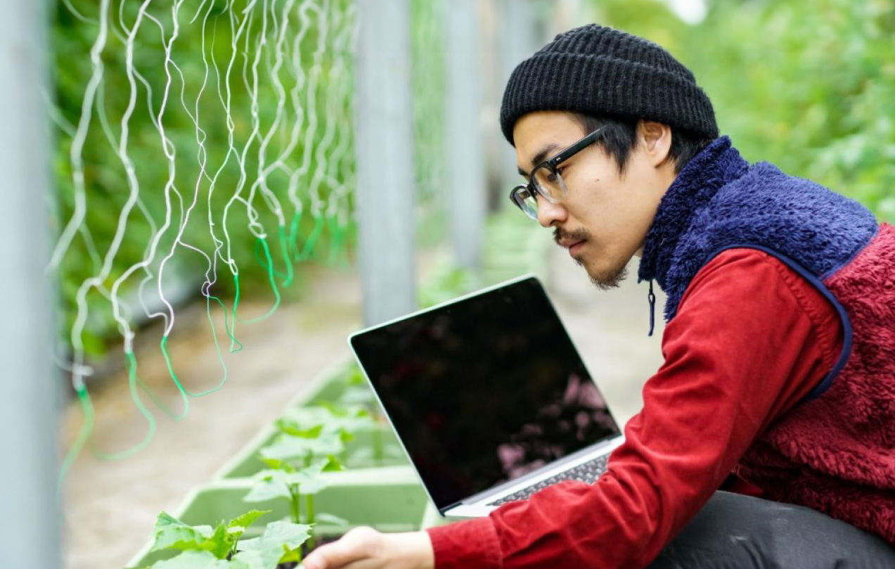 An RRU student squats down next to a row of plants holding his laptop