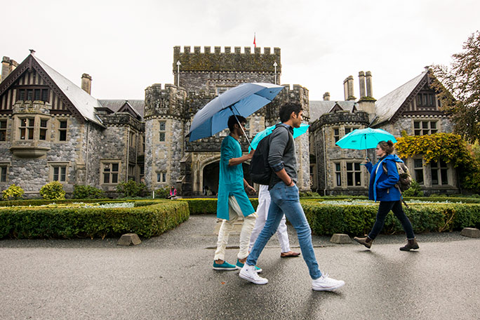 A group of international students holding umbrellas walk past Hatley Castle on RRU's Colwood Campus.