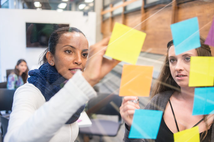 Two RRU students place colourful sticky notes to a glass wall while their classmates observe from their seats.