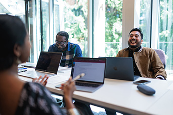 Three colleagues sit around a table in a meeting room, engaged in conversation, with a glass wall behind them covered in welcoming notes and drawings.