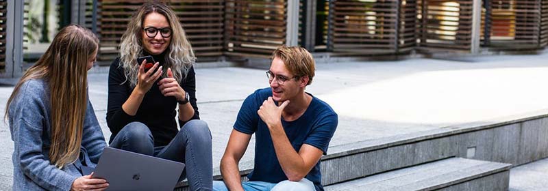 Three people sit closely on a sidewalk ledge with a laptop and a smart phone.