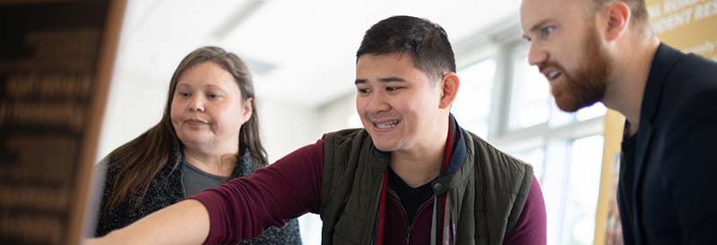 Three students lean in to examine a flipchart, where the middle person is pointing.