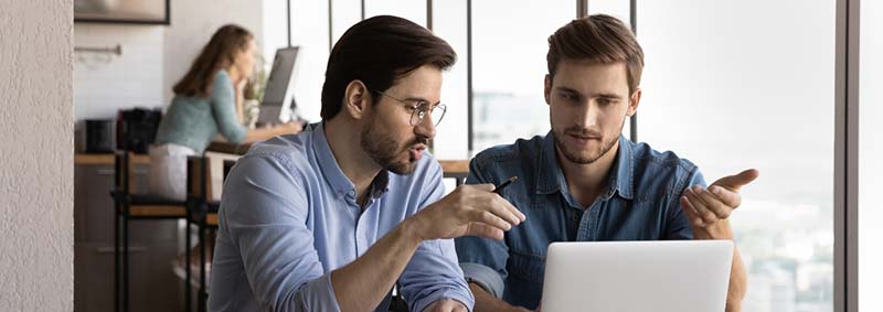 Two people sitting at a desk near a window discuss a laptop screen attentively.