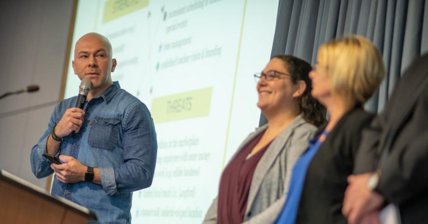 Speaker presenting at a podium next to participants at the Organizational Communication Challenge. Photo by Dan Anthon.