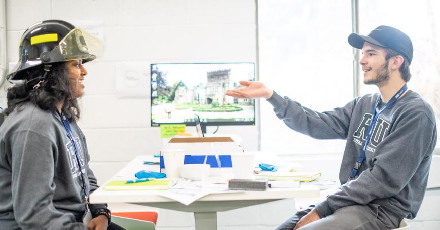 Two students in RRU hoodies have a discussion. One in a firefighters hat.