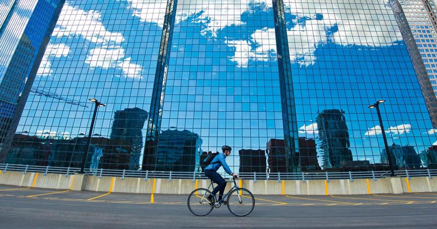 Person riding their bike on a highway next to a glass building reflecting a blue sky