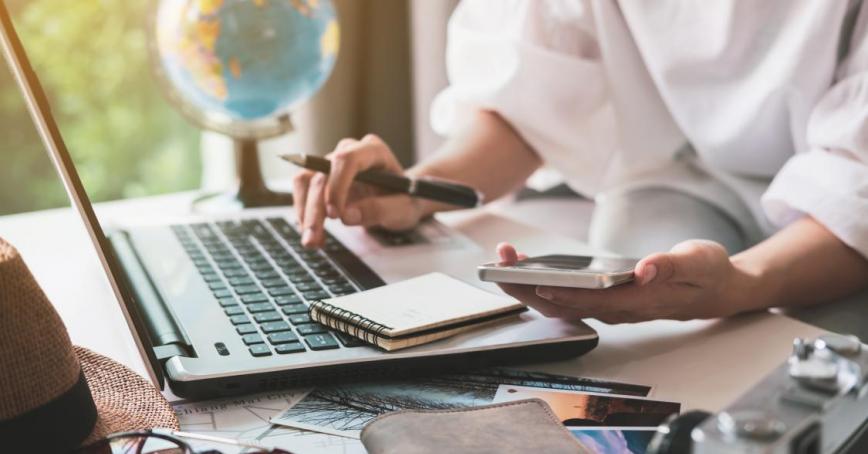 A person works on a laptop while holding a pen and a phone with a globe in the background.