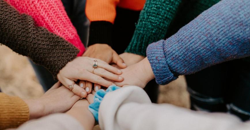 A circle of people in different colour sweaters with their hands piled in the centre. 