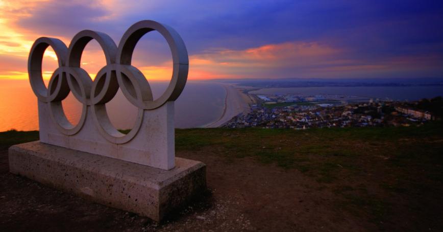 A statue of the Olympic rings atop a mountain overlooking a city by the ocean