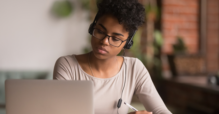 A woman wearing headphones takes notes while looking at a laptop display.