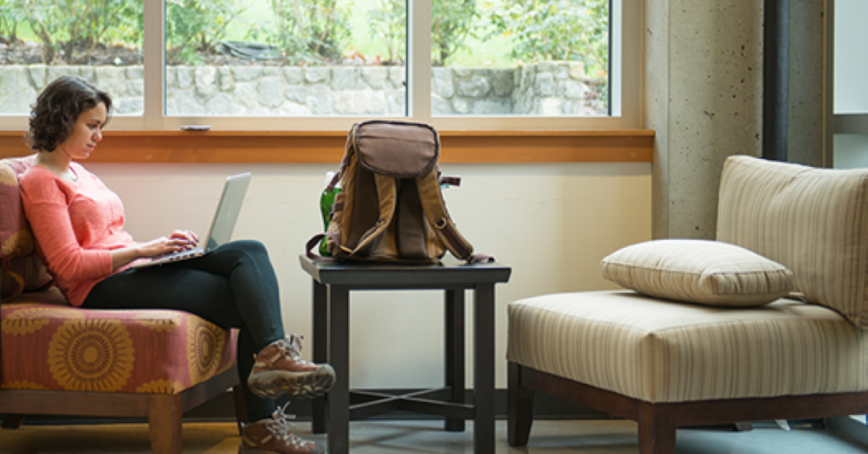 A woman in a coral coloured top and dark pants and hiking boots sits on a lounge chair to use a laptop. A backpack sits on a table nearby.