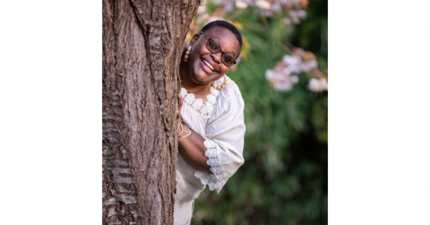 A smiling woman peeks out from behind a large tree, there is greenery in the background.