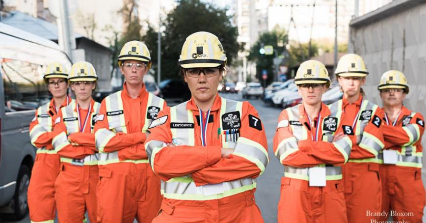 Group of seven women wearing hard hats and orange safety jumpsuits stand with their arms crossed -- they are a mine rescue team.