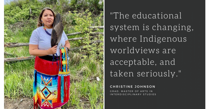 Christine Johnson stands in front of green trees and bushes. She is holding a full wing and is dressed in a bright red skirt and a beaded necklace.
