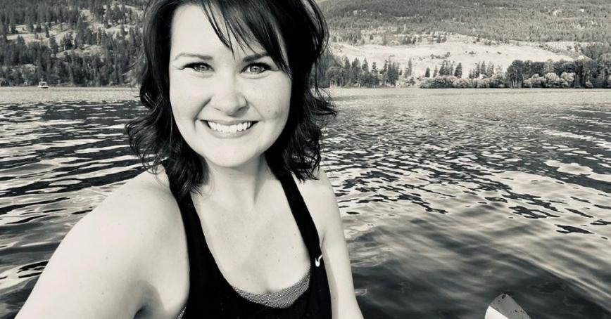 Black and white photo of a smiling woman with mid-length dark hair taken on a kayak from the middle of a lake. A treed and hilly landscape is in the background.