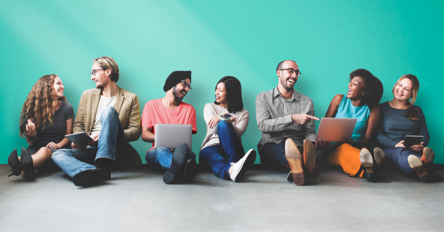 A group of seven workers of various ethnic backgrounds sit on the floor in front of a teal wall while tallking and laughing, some with laptops open.