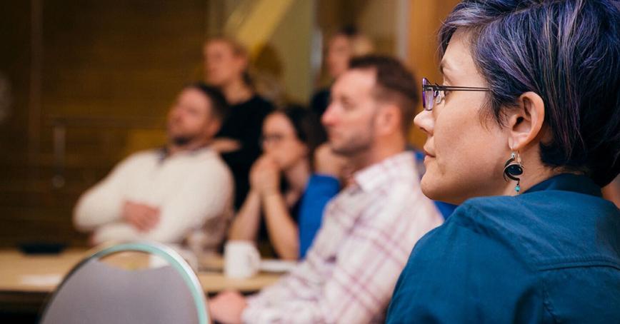 Prof. Jaigris Hodson studiously watches a presenter at a conference. Her purple hair glints in her glasses.